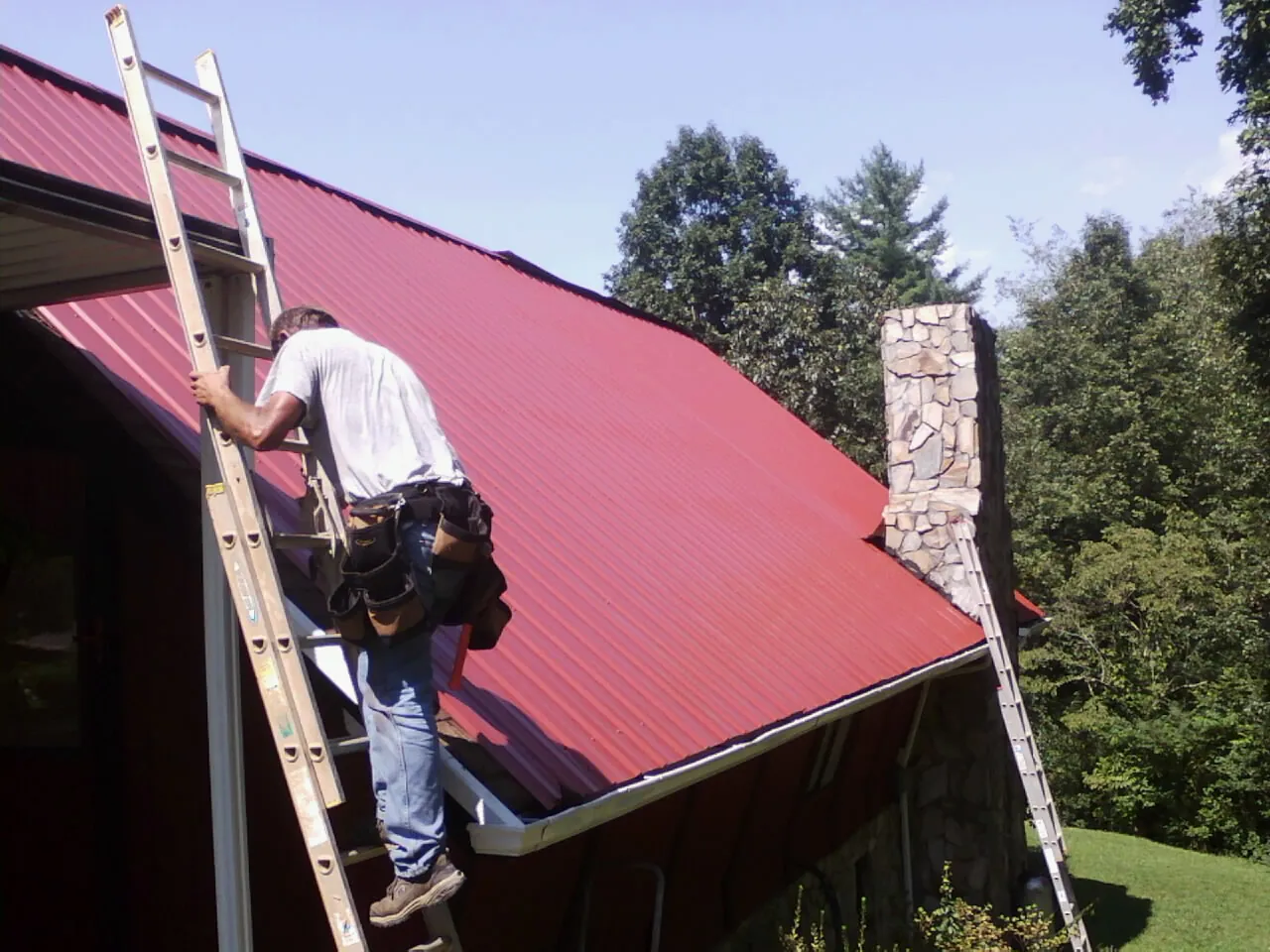 Roofer on a ladder beside a red metal roof and stone chimney — Soffit & Fascia Repair by Roofing Brothers in Brentwood